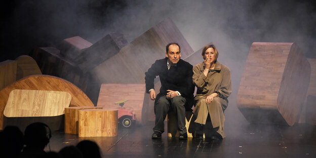 Eine Schauspielerin als Angela Merkel sitzt an einem Tisch und breitet die Arme aus Eine Schauspielerin als Angela Merkel sitzt an einem Tisch und breitet die Arme aus