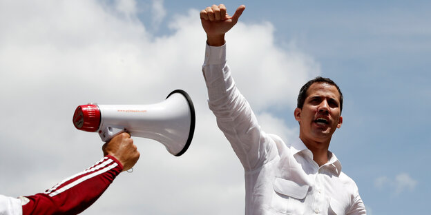 Guaidó in Sprecherpose gegen den blauen Himmel. In das Foto ragt ein Arm, die Hand hält ein Megaphon Guaidó in Sprecherpose gegen den blauen Himmel. In das Foto ragt ein Arm, die Hand hält ein Megaphon