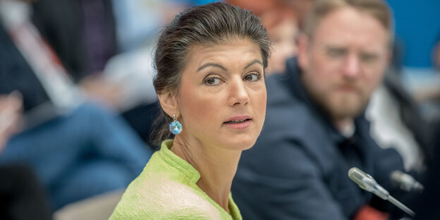 Sahra Wagenknecht sitzt vor einem Mikro inmitten mehrere Menschen Sahra Wagenknecht sitzt vor einem Mikro inmitten mehrere Menschen