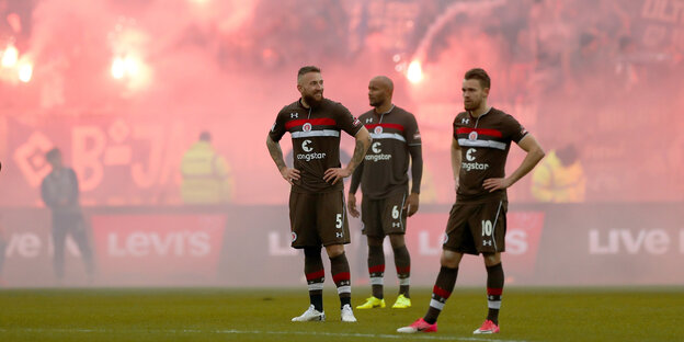 Drei Spieler des FC St. Pauli stehen auf dem Spielfeld des Millerntor-Stadions, im Hintergrund qualmen Bengalos. Drei Spieler des FC St. Pauli stehen auf dem Spielfeld des Millerntor-Stadions, im Hintergrund qualmen Bengalos.