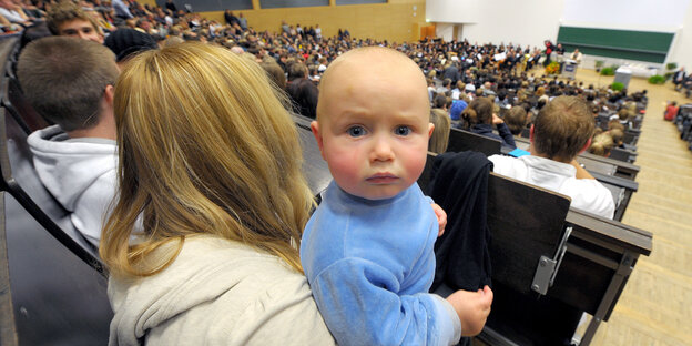 Eine Frau sitzt mit einem Baby auf dem Arm in einem Hörsaal. Eine Frau sitzt mit einem Baby auf dem Arm in einem Hörsaal.