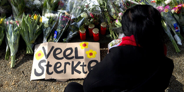 Eine Frau legt Blumen nieder. Auf dem Boden liegt ein Schild, auf dem auf Niederländisch "Viel Kraft" steht. Eine Frau legt Blumen nieder. Auf dem Boden liegt ein Schild, auf dem auf Niederländisch "Viel Kraft" steht.