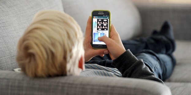 Ein Jugendlicher liegt auf einem Sofa und schaut auf sein Smartphone in seiner Hand Ein Jugendlicher liegt auf einem Sofa und schaut auf sein Smartphone in seiner Hand