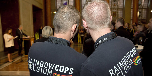 Zwei Männer sind vin hinten zu sehen, sie tragen T-Shirts mit der Aufschrift "Rainbow Cops Belgium" Zwei Männer sind vin hinten zu sehen, sie tragen T-Shirts mit der Aufschrift "Rainbow Cops Belgium"