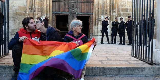 Drei Männer sitzen mit einer Regenbogenfahne vor einer Kirche im spanischen Bistum in Alcalá de Henares und protestieren gegen die Konversionstherapien. Drei Männer sitzen mit einer Regenbogenfahne vor einer Kirche im spanischen Bistum in Alcalá de Henares und protestieren gegen die Konversionstherapien.