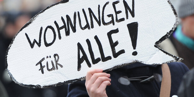 Eine Hand hält ein Schild in Form von einer Sprechblase hoch, darauf steht: "Wohnungen für alle!"