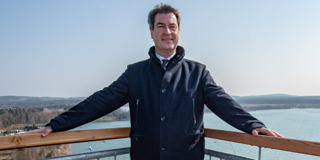 Markus Söder steht auf einem Balkon, hinter ihm ein See