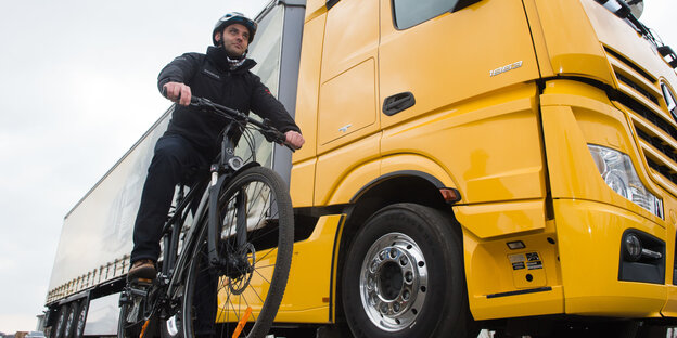 ein Radfahrer mit Helm wartet neben einem riesigen gelben Lkw ein Radfahrer mit Helm wartet neben einem riesigen gelben Lkw