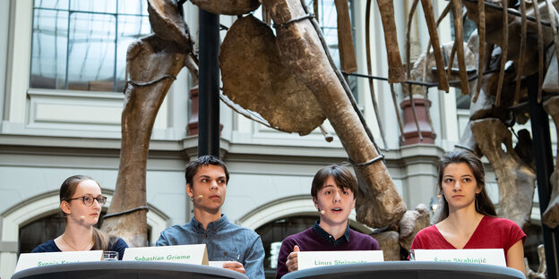 Junge Aktivist*innen sitzen vor einem Dinosaurierskelett auf einem Podium. Junge Aktivist*innen sitzen vor einem Dinosaurierskelett auf einem Podium.
