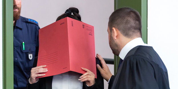 Eine Frau hält ein rotes Heft vor ihr Gesicht. Ein Mann in Anwaltsrobe berührt sie an der Schulter ubnd redet mit ihr. Eine Frau hält ein rotes Heft vor ihr Gesicht. Ein Mann in Anwaltsrobe berührt sie an der Schulter ubnd redet mit ihr.
