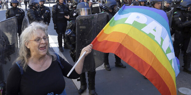 Eine FRau demonstriert mit Regenbogenflagge mit der Aufschrift „Paix“ („Frieden“), hinter ihr steht eine Polizei-Blockade Eine FRau demonstriert mit Regenbogenflagge mit der Aufschrift „Paix“ („Frieden“), hinter ihr steht eine Polizei-Blockade