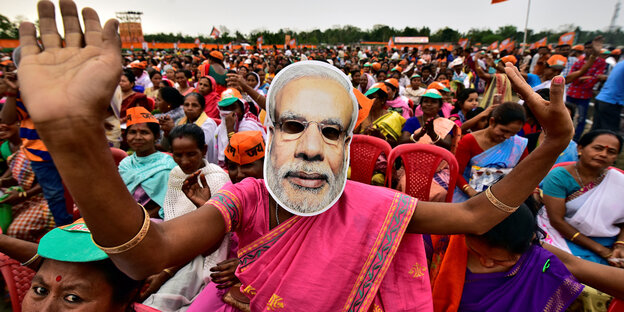 Eine Frau in pinkem Gewand steht in einer Menschenmenge und trägt eine Maske Narendra Modis Eine Frau in pinkem Gewand steht in einer Menschenmenge und trägt eine Maske Narendra Modis