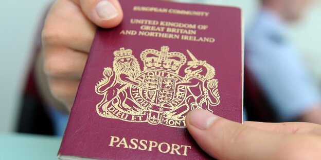 Eine Hand überreicht einer anderen Hand einen dunkelroten britischen Reisepass Eine Hand überreicht einer anderen Hand einen dunkelroten britischen Reisepass