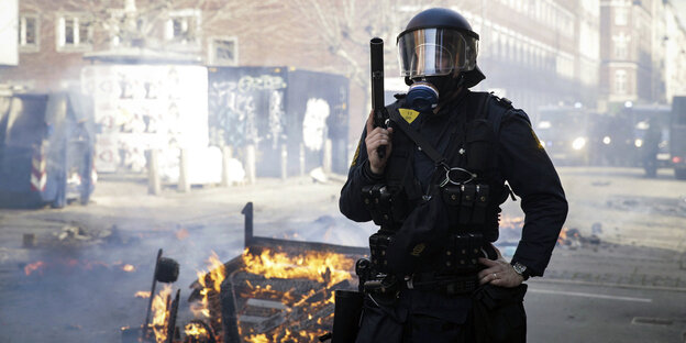 Ein Polizist in Riotuniform mit einer gezogenen Waffe vor einer kleinen brennenden Barrikade Ein Polizist in Riotuniform mit einer gezogenen Waffe vor einer kleinen brennenden Barrikade
