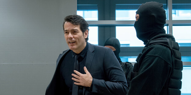 Adrian U. spricht, neben ihm zwei maskierte Polizisten Adrian U. spricht, neben ihm zwei maskierte Polizisten
