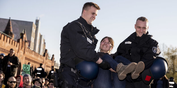 Zwei Polizisten tragen eine Klimaaktivist*in weg. Alle lächeln Zwei Polizisten tragen eine Klimaaktivist*in weg. Alle lächeln