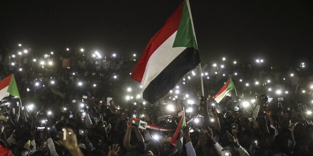 Menschen halten bei Nacht Smartphones in die Luft, in der Mitte des Bildes hält jemand die sudanesische Fahne hoch Menschen halten bei Nacht Smartphones in die Luft, in der Mitte des Bildes hält jemand die sudanesische Fahne hoch