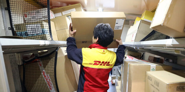 Eine DHL-Mitarbeiterin belädt ein Lieferfahrzeug mit Paketen. Sie ist von hinten zu sehen, wie sie ein Paket in einen Lieferraum mit vielen Paketen hebt. Eine DHL-Mitarbeiterin belädt ein Lieferfahrzeug mit Paketen. Sie ist von hinten zu sehen, wie sie ein Paket in einen Lieferraum mit vielen Paketen hebt.
