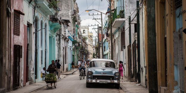 Eine Straßenszene in Havanna zeigt ein geparktes Auto, mehrere Fußgänger und bunte Häuser links und rechts