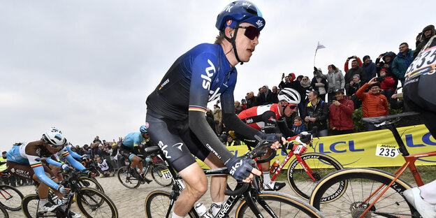 Ein Radsportler mit getönter Sonnenbrille ist im Vordergrund zu sehen, dicht hinter und vor ihm fahren weitere Radrennfahrer. Im Hintergrund stehen hinter einer Werbebande Zuschauer und Zuschauerinnen.