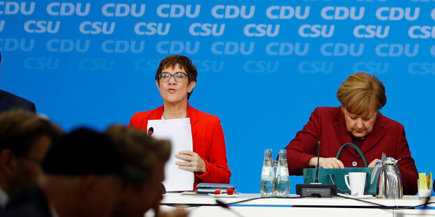 Annegret Kramp-Karrenbauer und Angela Merkel sitzen auf einem Podium nebeneinander. Kanzlerin Merkel sucht etwas in ihrer Handtasche Annegret Kramp-Karrenbauer und Angela Merkel sitzen auf einem Podium nebeneinander. Kanzlerin Merkel sucht etwas in ihrer Handtasche