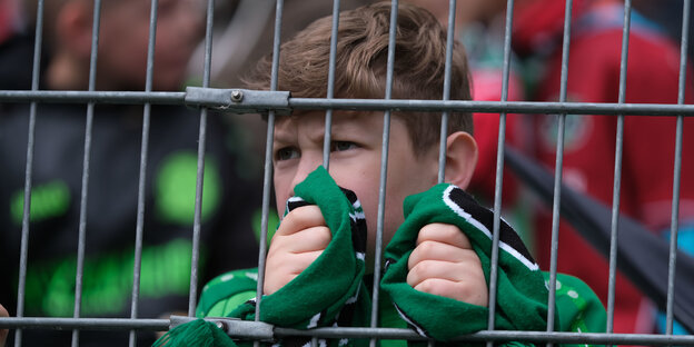 Ein kleiner Junge mit dem grünen Fan-Schal Hannovers steht hinter einem Zaun. Ein kleiner Junge mit dem grünen Fan-Schal Hannovers steht hinter einem Zaun.