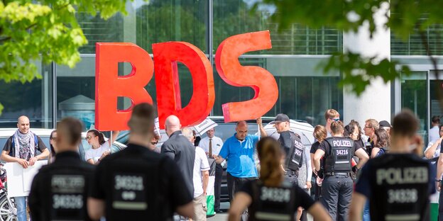 AktivistInnen halten riesige Buchstaben "BDS" hoch, davor stehen PolizistInnen AktivistInnen halten riesige Buchstaben "BDS" hoch, davor stehen PolizistInnen