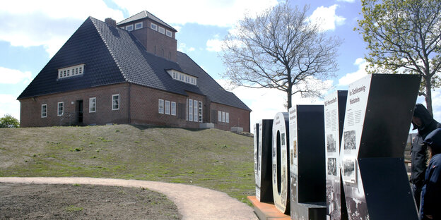 Halle mit tief herabgezogenem Walmdach auf einem Hügeln, im Vordergrund bildet eine Buchstabenskulptur das Wort Volk Halle mit tief herabgezogenem Walmdach auf einem Hügeln, im Vordergrund bildet eine Buchstabenskulptur das Wort Volk