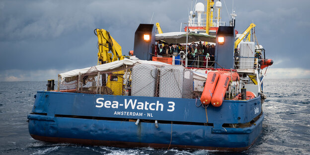 Auf dem Seenotrettungsschiff „Sea Watch 3“ stehen Gerettete und Crewmitglieder und winken. Auf dem Seenotrettungsschiff „Sea Watch 3“ stehen Gerettete und Crewmitglieder und winken.