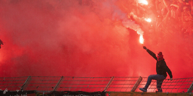 Ein Mann sitzt auf einem Stadionzaun und schwenkt eine bengalische Fackel Ein Mann sitzt auf einem Stadionzaun und schwenkt eine bengalische Fackel