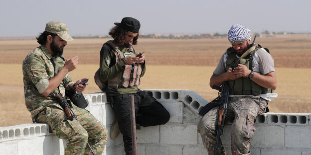 Drei Kämpfer mit Waffen sitzen auf einer Mauer und schauen in ihre Smartphones.