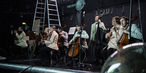 Musikerinnen und Musiker sitzen und stehen auf der Bühne im Residenztheater in München „Olympiapark in the Dark“ von Thom Luz. Sie spielen Geigen, Cellos und einen Bass.