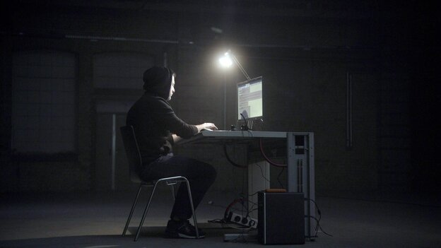 Ein dunkler Raum, eine Person im Hoodie sitzt vor einem Computer