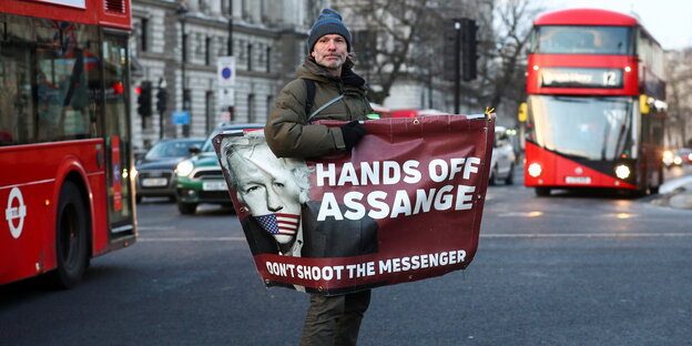Mann auf einer Straße in London, man sieht zwei rote Doppeldeckerbusse. In den Händen hält er ein Transparent: "Hands off Assange"