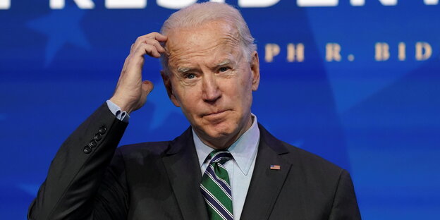 Joe Biden steht vor blauem Hintergrund und kratzt sich nachdenklich am Kopf