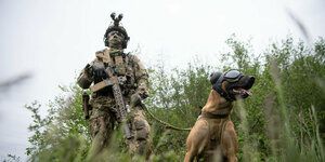 Ein Kommandosoldat des Kommando Spezialkräfte (KSK) der Bundeswehr, steht während eines Videodrehs zum Tag der Bundeswehr mit einem Zugriffsdiensthund, der einen Augen- und Ohrenschutz trägt, in einer Wiese.