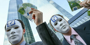 Zwei Männer mit weißen masken stehen vor den Bürotürmen der Deutschen Bank in Frankfurt und halten falsche Geldschein in den Händen