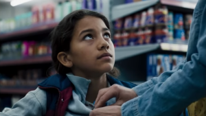 Standbild aus dem Trailer von „Platzspitzbaby“. Einem Mädchen mit langen Haaren wird im Supermarkt ein Produkt unter die Jacke geschoben. Sie schaut nach oben.