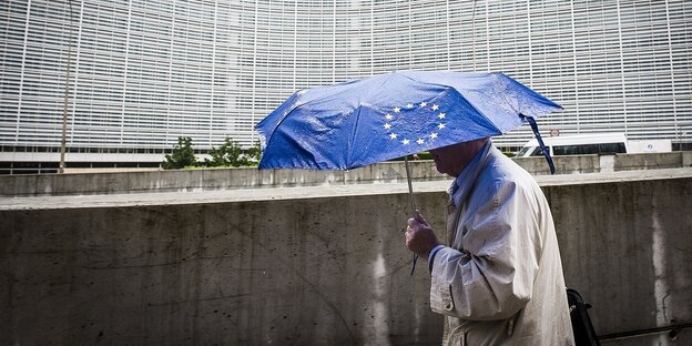Ein Mann trägt eine blauen Schirm mit dem Logo der EU und geht am Gebäude der Europäischen KOmmission entlang