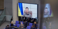 Wolodimir Selenski ist auf einem Videobildschirm zu sehen, vor ihm sitzen Bundestags-Abegeordnete und Minister:innen mit Kopfhörern