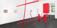 Sicht in die Ausstellung "Digestion" von Henrik Olesen. Im Vordergrund ist die rot lackierte, abstrakte Metallskulptur "Monument after Anthony Caro" zu sehen: Metallstreben überkreutzen sich waagerecht und senkrecht. Dahinter hängen von links nach rechts