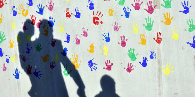 Drei Schatten zeichnen sich auf einer Wand voller Abdrücke von Kinderhänden ab. Drei Schatten zeichnen sich auf einer Wand voller Abdrücke von Kinderhänden ab.