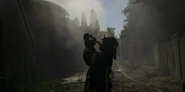 Ein Feuerwehrmann in voller Montur nimmt einen tiefen Schluck aus einer Trinkflasche, hinter ihm ein schwelendes Gebäude