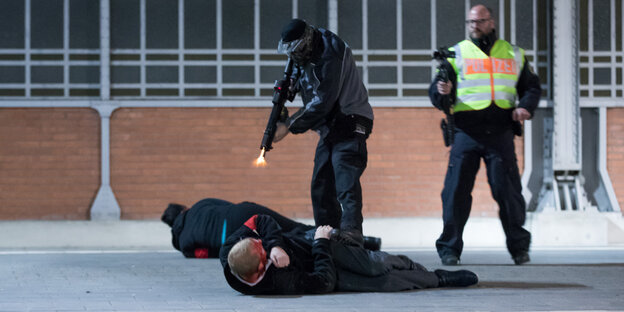 Zwei Menschen liegen auf einem Bahnsteig. Auf einen von ihnen tritt ein maskierter Polizist und hält in Richtung von dessen rot gefärbtem Kopf ein Gewehr, aus dem Mündungsfeuer zu kommen scheint. Im Hintergund filmt ein weiterer Polizist die Szene.