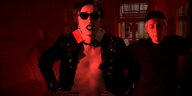 Videostill aus "Becoming Rabe": In rotes Licht getaucht schauen zwei Menschen in die Kamera. Die Person im Zemtrum trägt eine Sonnenbrille und hält die Hände in die Hüften gestützt. Sie trägt eine Lederjacke über dem nackten Oberkörper. Sie trägt Kreitkar