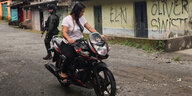 Eine Motorradfahrerin steht an einer Straßenecke, im Hintergrund eine Hauswand mit mehreren Graffiti-Schriftzügen