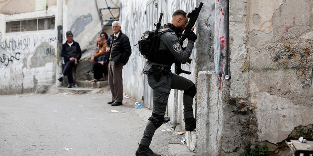 Ein israelischer Soldat mit Maschinengewehr betritt ein Haus. Im Hintergrund stehen ältere Passanten, die ihn argwöhnlisch beobachten