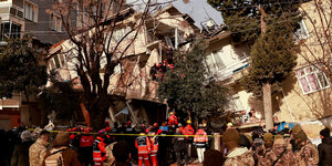 Feuerwehrleute und andere Uniformierte stehen vor einem zum großen Teil eingestürzten Haus