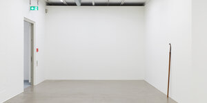 Ein fast leerer Ausstellungsraum, rechts lehnt eine Hippe an der Wand