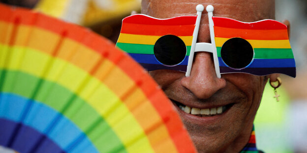 Ein Mann mit einer Regenbogenbrille und einem Regenbogenfächer lächelt in die Kamera